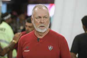 Mano Menezes, técnico do Fluminense. Ele tem cabelo e barba grisalhos e olhos verdes. Usa uma camisa na cor vermelha com o escudo do Fluminense no lado esquerdo do peito. O escudo do Fluminense, por sua vez, é tricolor (grená, branco e verde), com as siglas 'FFC' em fonte gótica dentro do emblema.